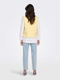 Geel gebreid vest over een wit overhemd, gecombineerd met lichtblauwe spijkerbroek en grijze sandalen met hak. Gladde textuur en een ontspannen pasvorm.