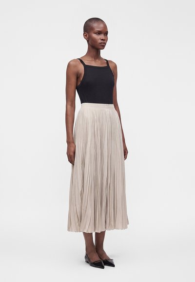 Femme portant un haut sans manches noir, une jupe midi plissée beige et des ballerines pointues noires, debout devant un fond clair uni.