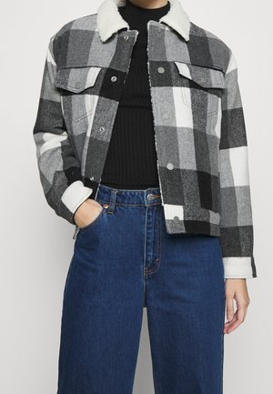 Person iført en sort og hvid ternet jakke med hvid krave, sort ribstrikket sweater og højtaljede blå jeans, hånd i lommen.