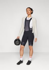 Sorte sykkelbuks med ribbet tekstur, hvit topp med mesh, lys beige langermet cardigan, holder en svart hjelm. Hvite og røde stripete sokker.