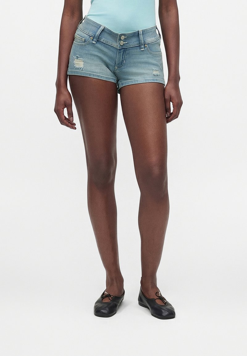 Άτομο με λεπτά πόδια που φοράει ανοιχτά μπλε denim σορτς με φθαρμένες λεπτομέρειες, ανοιχτό μπλε τοπ και μαύρα μπαλαρίνια, στέκεται μπροστά σε απλό φόντο.