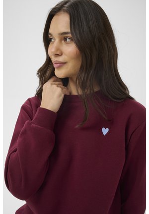 Kvinde med langt mørkt hår iført en burgunderrød sweatshirt med et lille lyseblåt hjerte på brystet, der kigger til siden.
