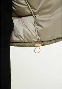 Detalj av puffjacka visar ett glänsande beige innerfoder, svart mudd och en guldfärgad hårdvarukrok för stängning i nederkant.