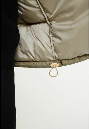 Detalj av puffjacka visar ett glänsande beige innerfoder, svart mudd och en guldfärgad hårdvarukrok för stängning i nederkant.