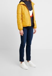 Donna in piedi che indossa una giacca puffer gialla senape, un hoodie a blocchi di colore blu navy, jeans blu scuro e sneakers bianche su uno sfondo neutro.
