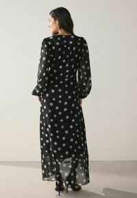 Rochie lungă neagră cu mâneci lungi, cu un model spiral alb, din material transparent și siluetă fluidă. Prezintă un tiv stratificat și cute la umeri.