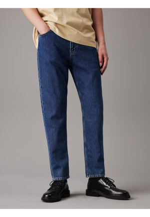 Persoon draagt blauwe jeans met rechte pijpen, beige t-shirt en zwarte leren veterschoenen, staand met één hand in de zak.