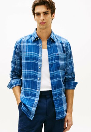 Ung mand med krøllet brunt hår iført en blå ternet skjorte over en hvid tanktop og mørkeblå bukser, stående med hænderne i lommerne.