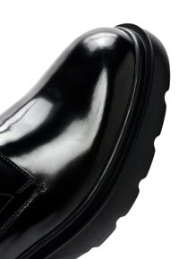 Zapato de cuero negro brillante con punta redondeada, que presenta una suela negra gruesa y texturizada y costuras visibles en la parte superior.