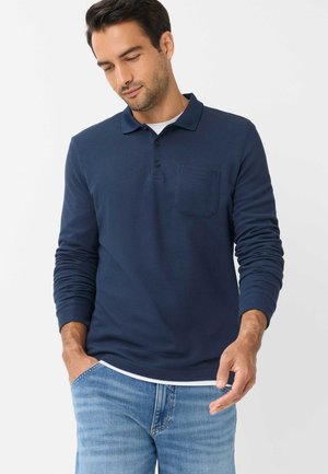 Man draagt een marineblauw poloshirt met lange mouwen over een wit hemd en lichtblauwe spijkerbroek, met één hand in de zak.