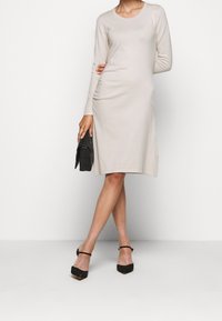 Krešlové béžové šaty po kolena s dlouhými rukávy, kulatým výstřihem, texturovanou látkou a bočními řasenými detaily. Nosí se s černými podpatky a malou černou kabelkou.