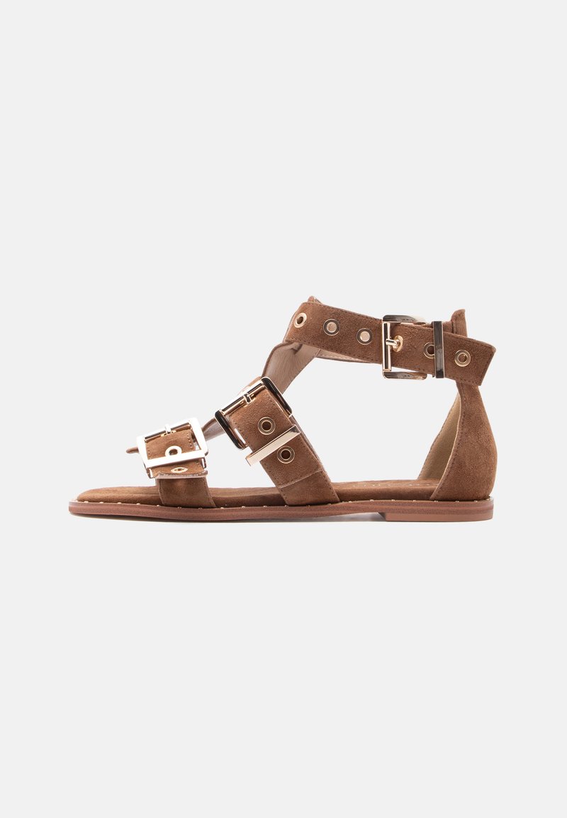 Καφέ σουέντ φλατ σανδάλι με τρεις ρυθμιζόμενες λουρίδες με αγκράφες και μεταλλικές τρύπες, ανοιχτό δάχτυλο και τακούνι, σε λευκό φόντο.