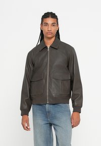 Nuori mies letitetyillä hiuksilla, päällä tummanruskea vetoketjullinen takki, jossa on etutaskut, ja päällä vaaleansiniset farkut, seisoo yksinkertaista valkoista taustaa vasten.