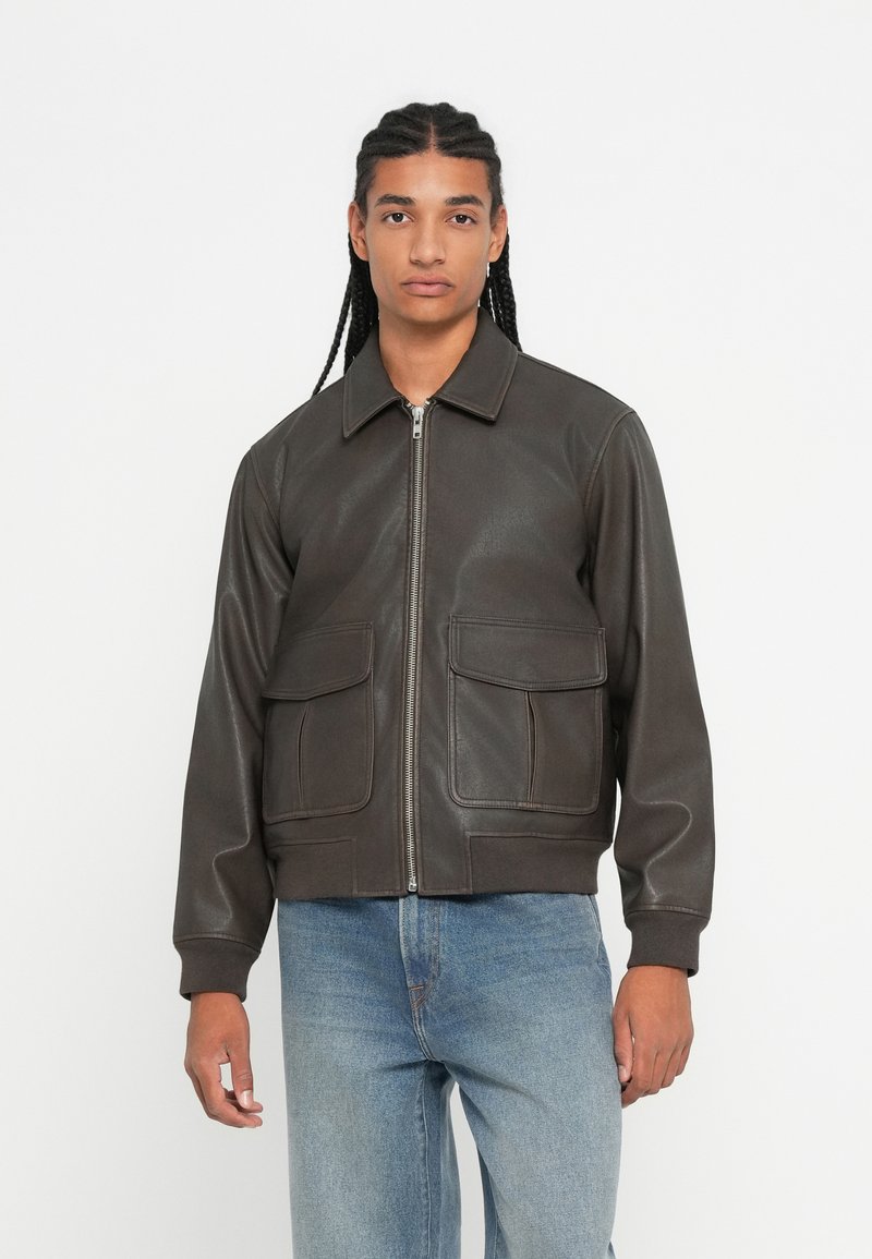 Nuori mies letitetyillä hiuksilla, päällä tummanruskea vetoketjullinen takki, jossa on etutaskut, ja päällä vaaleansiniset farkut, seisoo yksinkertaista valkoista taustaa vasten.