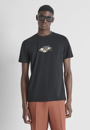 Giovane uomo con t-shirt nera con grafica, grandi occhiali da sole neri e pantaloni color ruggine su uno sfondo chiaro e uniforme.