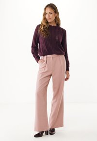 Donna in piedi con una mano in tasca, indossa un maglione a maglia viola, pantaloni larghi rosa chiaro e stivali con tacco scuro su sfondo bianco.