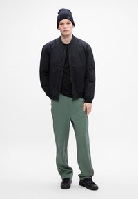 Schwarze Bomberjacke mit Reißverschluss, enganliegenden Bündchen und Seitentaschen, kombiniert mit hellgrünen gemusterten Jogginghosen und schwarzen Sneakers.