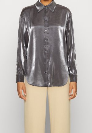 Kvinde iført en metallisk sølvfarvet langærmet skjorte med knapper kombineret med beige bukser med vide ben.