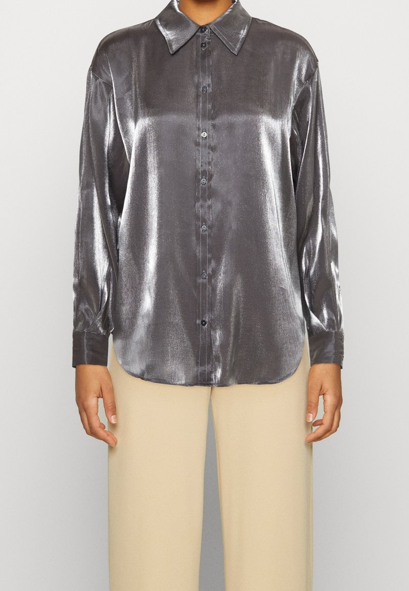 Femme portant une chemise argentée métallique à manches longues boutonnée, associée à un pantalon beige à jambes larges.
