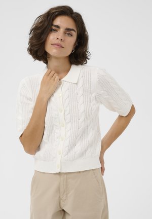 Kvinde med kort krøllet hår iført en hvid kabelstrikket kortærmet cardigan med knapper og beige bukser foran en ensfarvet baggrund.