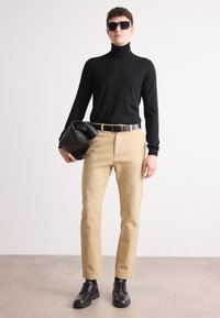 Pulover negru cu guler înalt, pantaloni bej, curea neagră și pantofi negri. Modelul ține o geantă tote neagră. Design minimalist, culori uni.