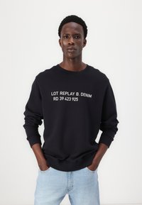 Fekete pulóver fehér felirattal, amelyen az "LOT REPLAY B. DENIM RD 39 423 925" olvasható. Kerek nyakú és bordás mandzsettájú, világoskék farmernadrággal párosítva.