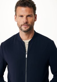 Marinblå bomberjacka med dragkedja, tillverkad av ett smidigt tyg. Har en rund halslinning och en slim fit. Bärs över en randig skjorta.