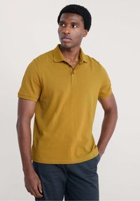 Sennepsgul polo-shirt med krave, korte ærmer og tre-knaps lukning. Fremstillet af blødt, struktureret stof.