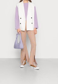 Gilet bianco doppiopetto, maglione testurizzato lavanda, pantaloni a quadri arancioni e bianchi, profonde aperture, borsa lavanda, sneakers marroni.