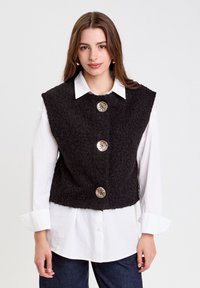 Schwarze, strukturiertes ärmelloses Oberteil im cropped Design, versehen mit vier großen silbernen Knöpfen, getragen über einem weißen Button-Down-Hemd.