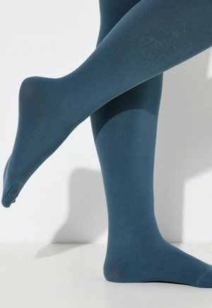 Due gambe incrociate che indossano collant opachi di colore blu teal su uno sfondo bianco semplice con ombre morbide.