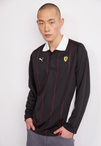 Juodas ilgomis rankovėmis polo marškinėliai su raudonomis juostelėmis, balta apykakle, Puma logotipas ant krūtinės ir Ferrari emblema. Tekstūruota medžiaga, laisvas siluetas.