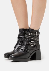 Botas de cuero negro hasta el tobillo con un tacón grueso, que presentan múltiples correas con tachuelas y un acento de hebilla alrededor del tobillo. Textura suave.