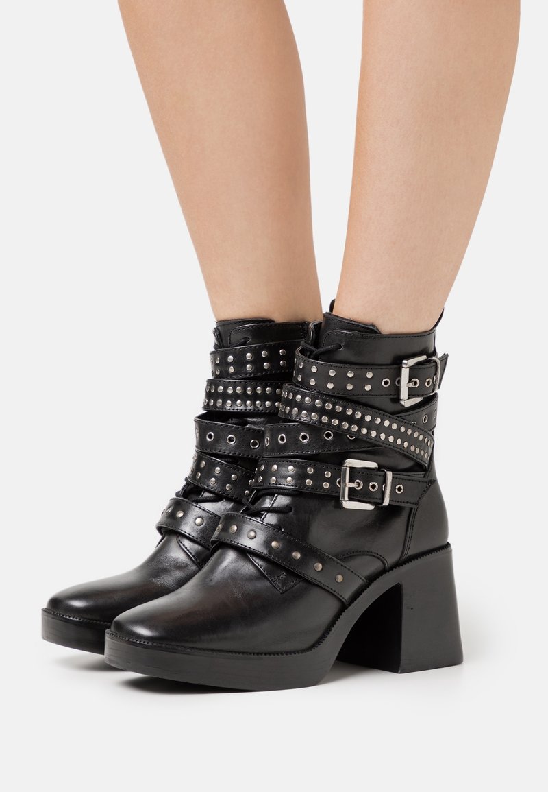 Botas de cuero negro hasta el tobillo con un tacón grueso, que presentan múltiples correas con tachuelas y un acento de hebilla alrededor del tobillo. Textura suave.