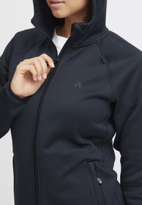 Giacca nera con cappuccio realizzata in tessuto morbido ed elastico, con zip frontale, tasche laterali e un logo discreto sul petto.