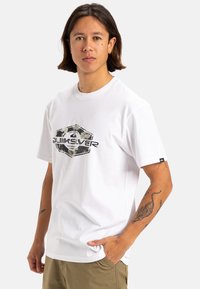 Camiseta de algodón blanca con un logo central negro y floral. Mangas cortas, cuello redondo, con sutiles acentos texturizados y una etiqueta.