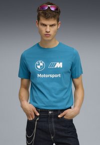 Turkio spalvos medvilnės marškinėliai su BMW ir M Motorsport logotipais balta spalva, derinami su tamsaus džinsinio audinio kelnėmis ir sidabriniu grandinės akcentu.