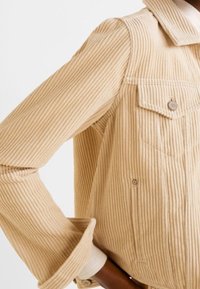 Beige corduroy jack met verticale ribbeltextuur, grote kraag, knoopzak op de borst en opgerolde mouwdetails.