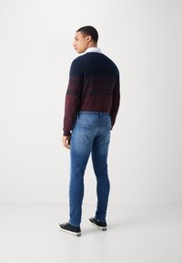 Suéter de punto para hombre en raya navy oscura y burdeos, combinado con jeans ajustados azules. Presenta mangas largas y zapatillas negras.