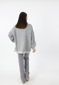 Oversized grijze trui met textuurgebreide stof, wijde mouwen en een gestreepte zoom. Gecombineerd met grijze flare-broeken. Effen witte achtergrond.
