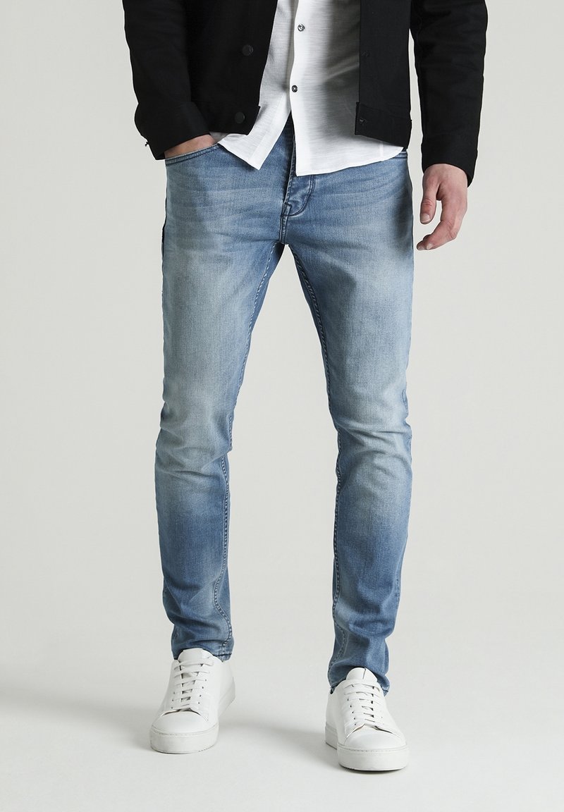 Homme portant un jean slim bleu clair, des baskets blanches, une chemise blanche et une veste noire, debout avec une main dans la poche.