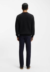 Schwarzer Pullover mit strukturierter Stoffqualität, geripptem Kragen und Bündchen, kombiniert mit dunklen Denim-Jeans, von hinten gesehen. Lässiges Schuhwerk vervollständigt den Look.