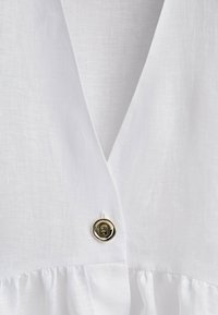 Vêtement en lin blanc avec un décolleté en V profond et un seul bouton rond argenté brillant près de la couture centrale au-dessus du tissu froncé.
