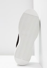 Semelle de chaussure blanche avec un motif de bande de roulement en zigzag, positionnée à la verticale contre l'angle des murs et du sol blancs.