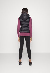 Svart pufferväst med huva, har en quiltad textur. Bärs över en lila långärmad topp, kombinerad med svarta leggings och sportskor.