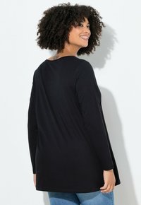 Schwarzes Langarmshirt mit geschwungenem Saum, runder Halslinie und weichem Material. Das Modell trägt es mit blauen Jeans und zeigt die Rückansicht.