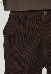 Pantalons en velours côtelé marron avec texture à côtes verticales, fermeture à bouton et taille structurée, montrés avec une partie d'un haut au-dessus.