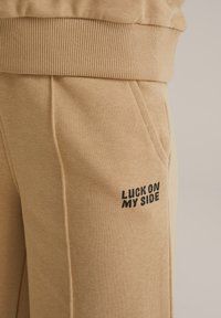 Beige jogger-housut, jotka on valmistettu pehmeästä puuvillasekoitteesta. Niissä on ribbikuvioitu vyötärö, sivutaskut ja musta "LUCK ON MY SIDE" -painatus.