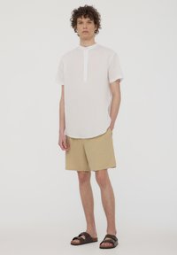 Camicetta bianca a maniche corte con chiusura a bottoni singoli, pantaloni beige e sandali scuri. La camicetta ha un orlo leggermente curvato.