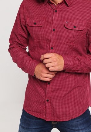 Hombre con camisa de cuadros roja con dos bolsillos en el pecho, ajustando el puño con ambas manos, vistiendo vaqueros azul oscuro.